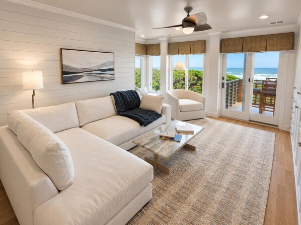 Un salon moderne avec un canapé blanc, une table basse, un ventilateur de plafond et une vue sur l’océan grâce à de grandes fenêtres.