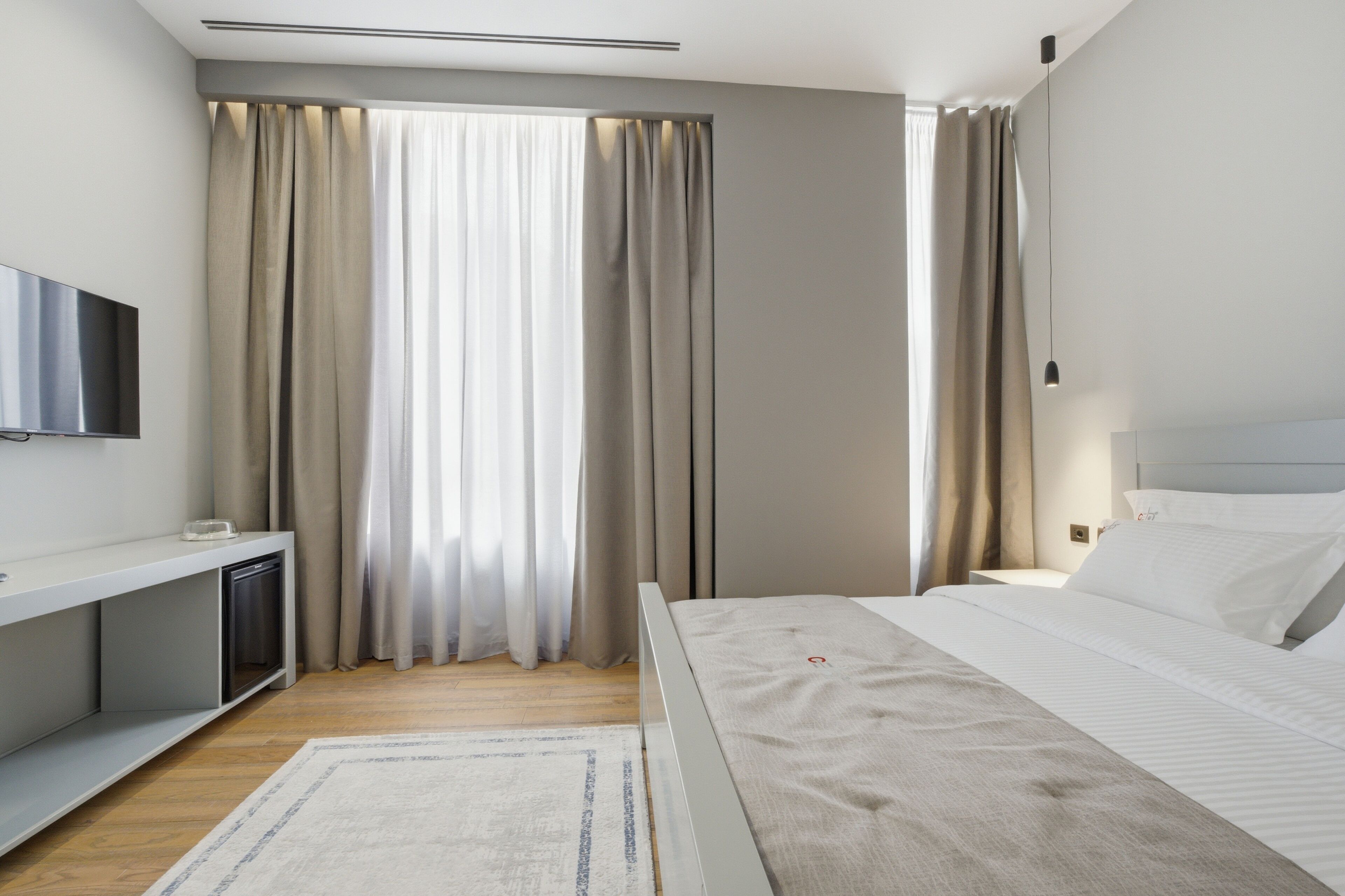 A modern hotel room with a bed, a TV, a desk, and a window with curtains.