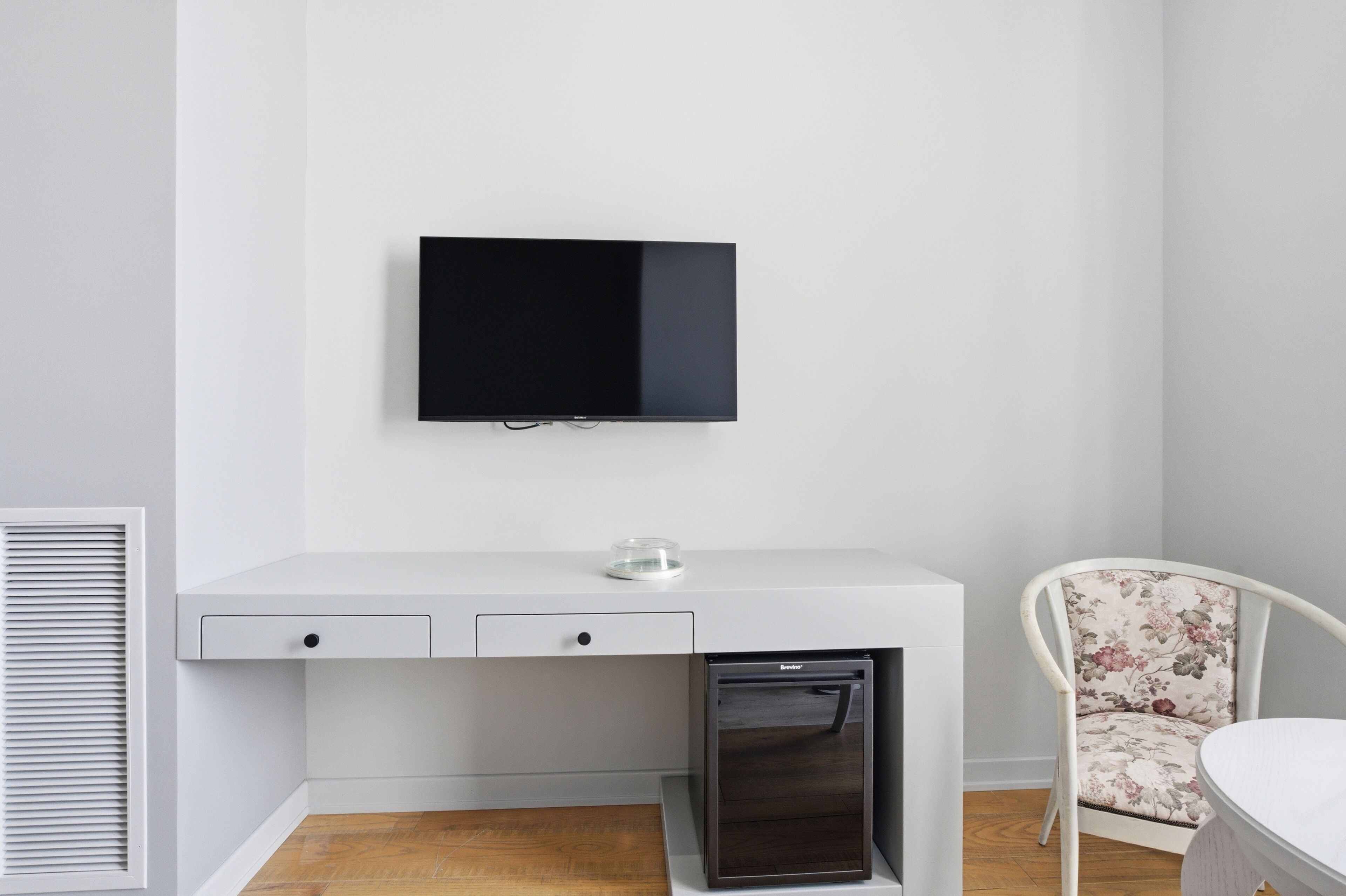 A white desk with a flat-screen TV, a small refrigerator, and a floral chair.