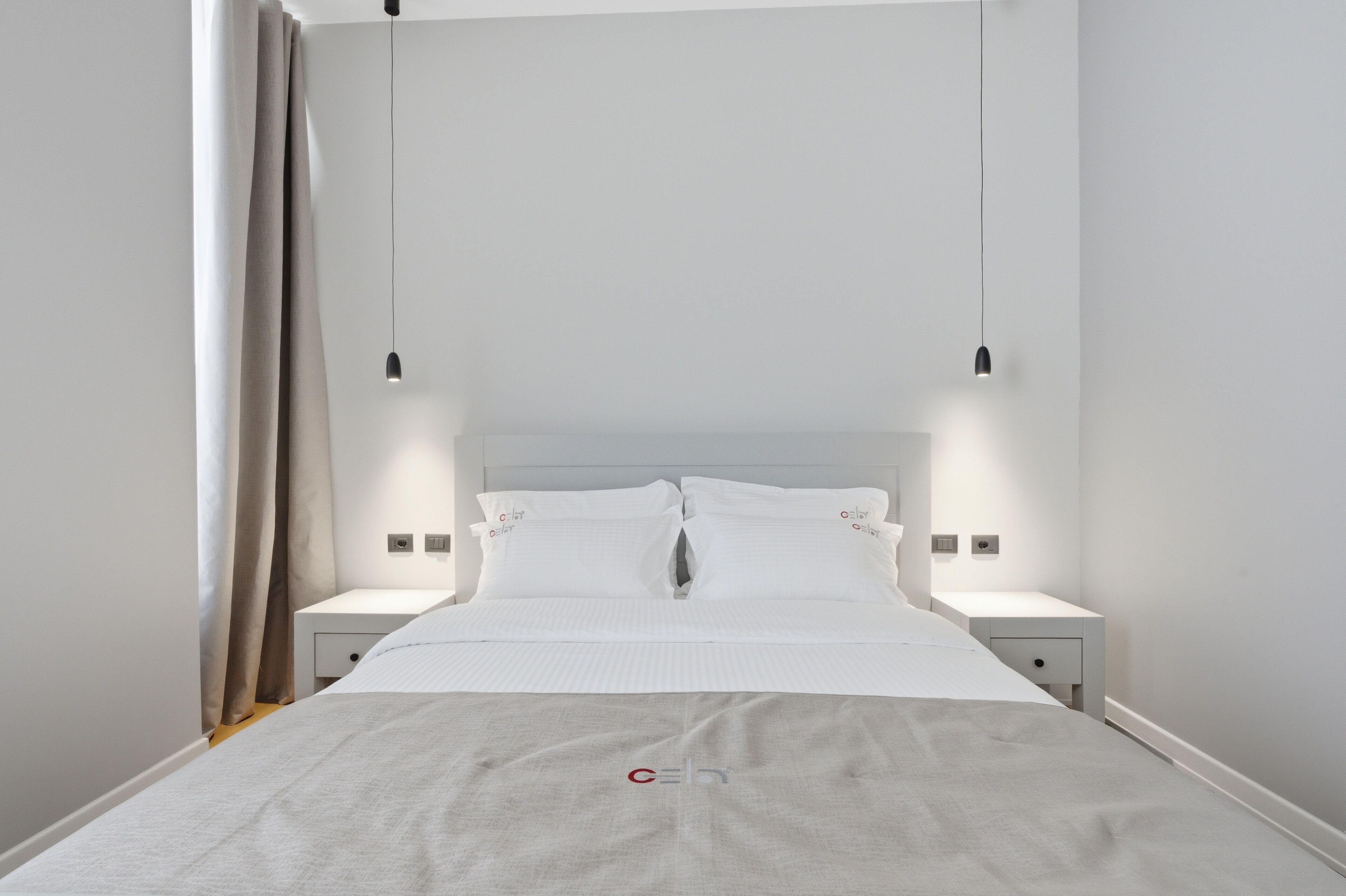 A modern hotel room with a bed, two bedside tables, a nightstand, and a hanging light fixture.