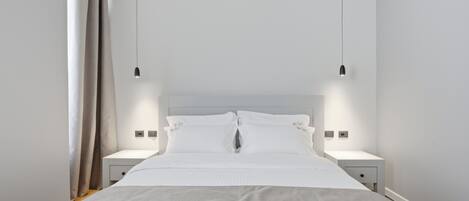A modern hotel room with a bed, two bedside tables, a nightstand, and a hanging light fixture.