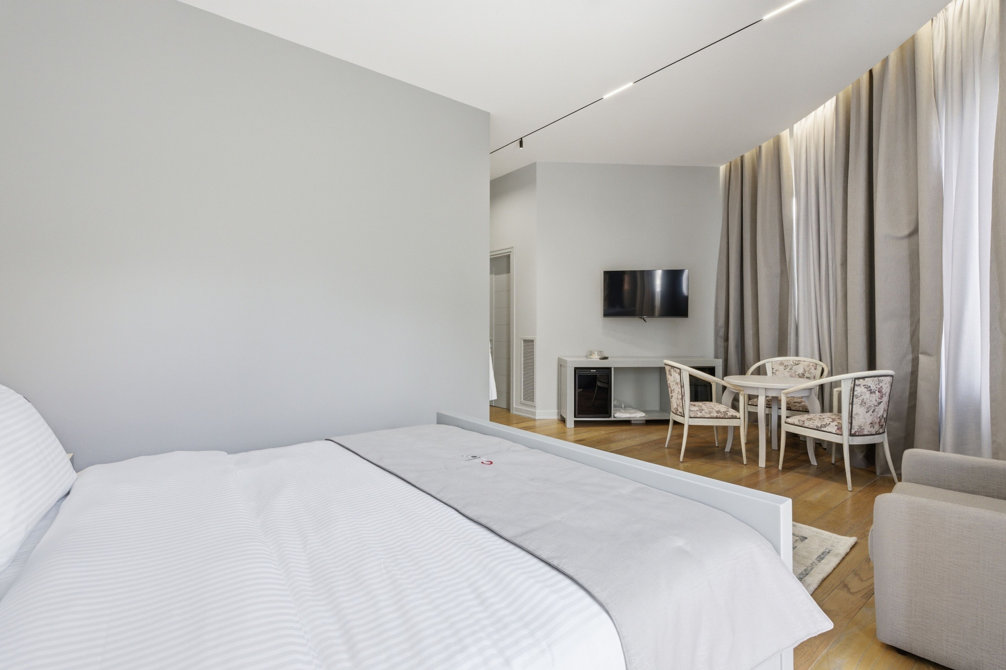 A modern hotel room with a large bed, a small table with chairs, a TV mounted on the wall, and a window with curtains.