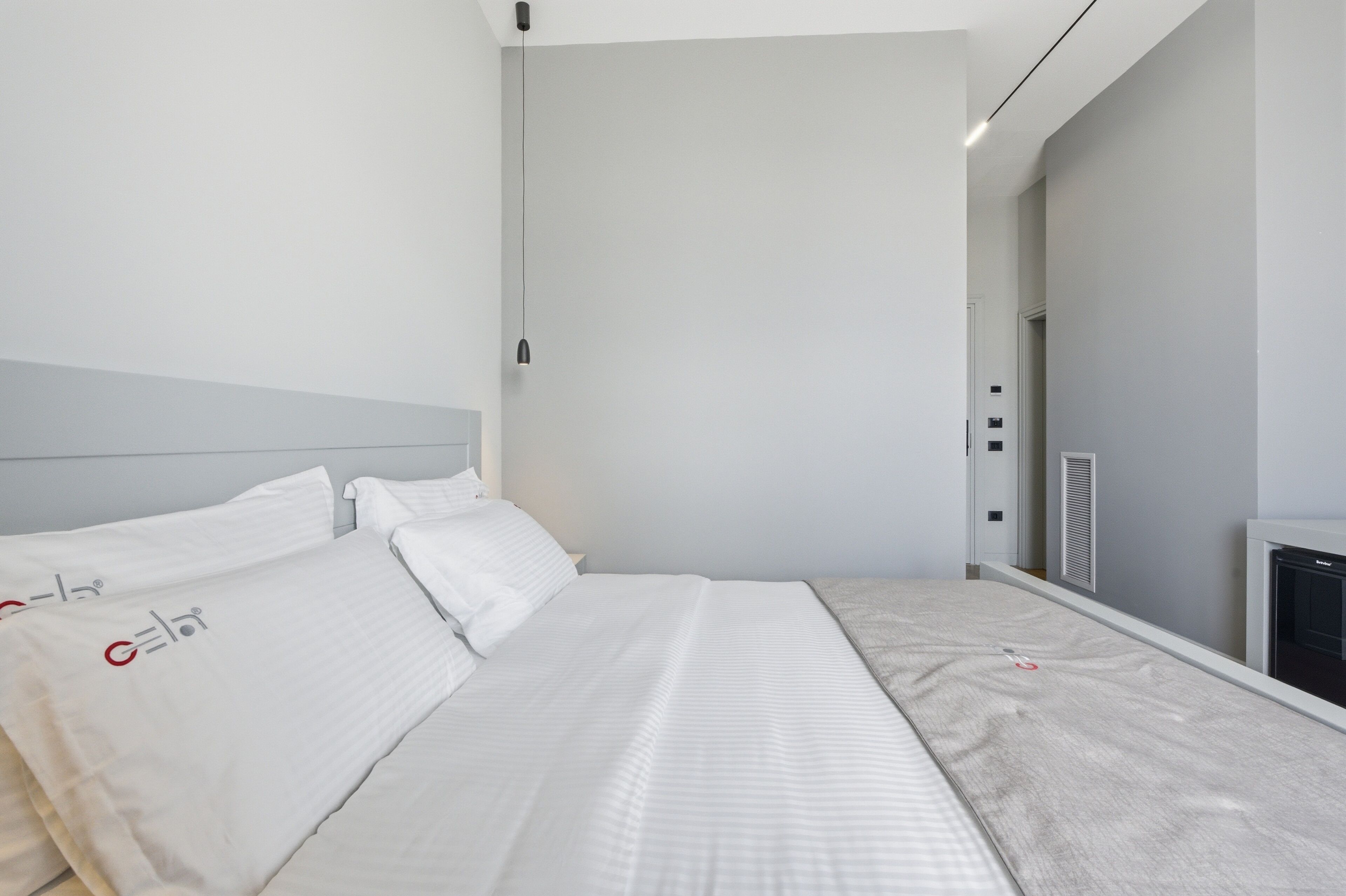 A modern hotel room with a large bed, a bedside table, a wall-mounted heater, and a door leading to a bathroom.