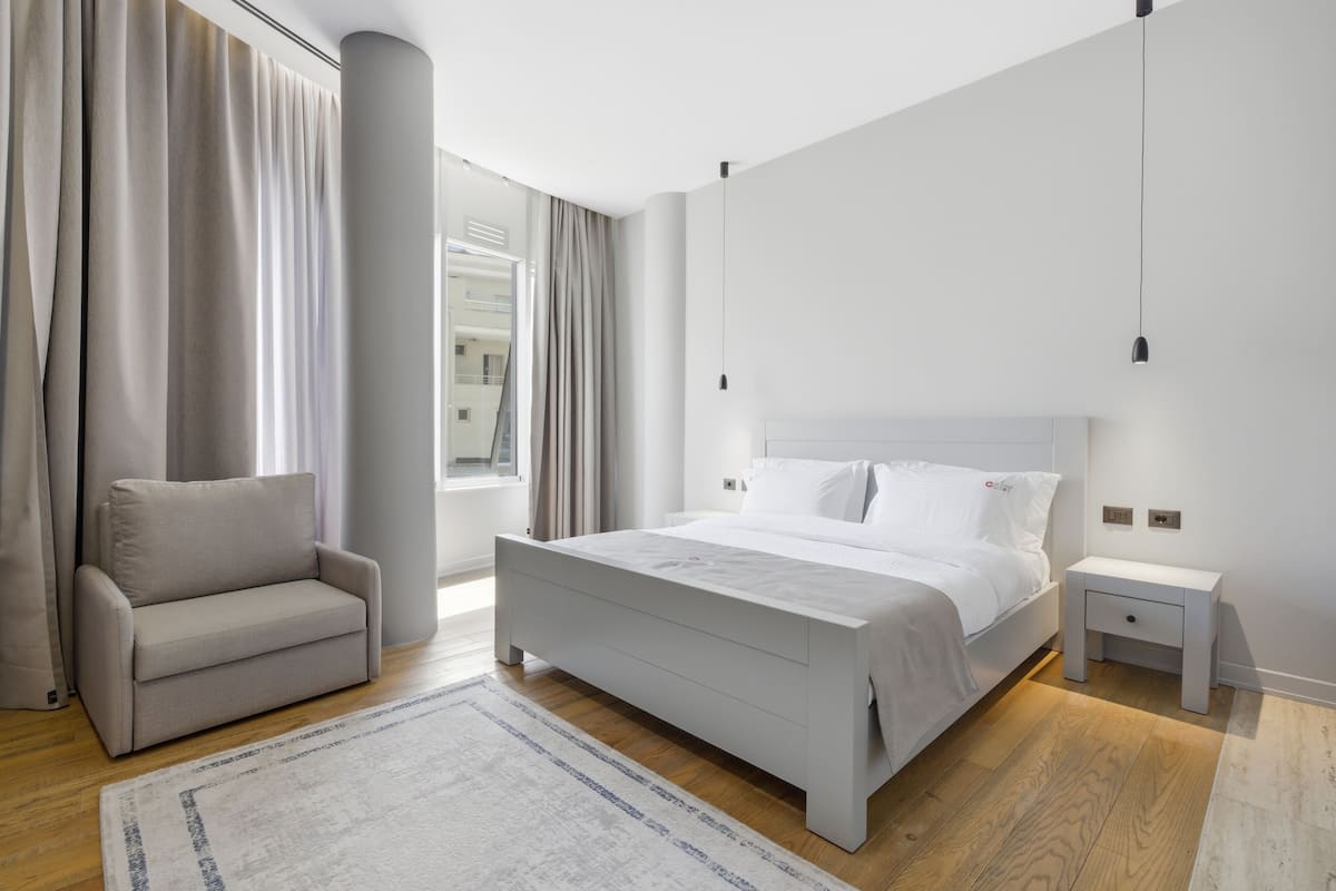 Un dormitorio moderno con cama, mesita de noche, sofá y un amplio ventanal con cortinas.