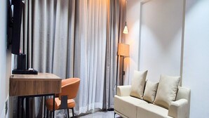 Une chambre d’hôtel moderne avec une table à manger en bois, des chaises orange, un canapé beige et un téléviseur fixé au mur.