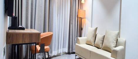 A modern hotel room with a wooden dining table, orange chairs, a beige sofa, and a TV mounted on the wall.