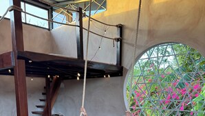 A person relaxing on a swing suspended from a wooden beam, with a view of pink flowers through a circular window. - Personal Retreat · Soaking Pool · Food Forest (Ometepe, Santa Cruz)