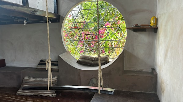 A hammock suspended by ropes, a window with a view of greenery, and a shelf with a yellow object.