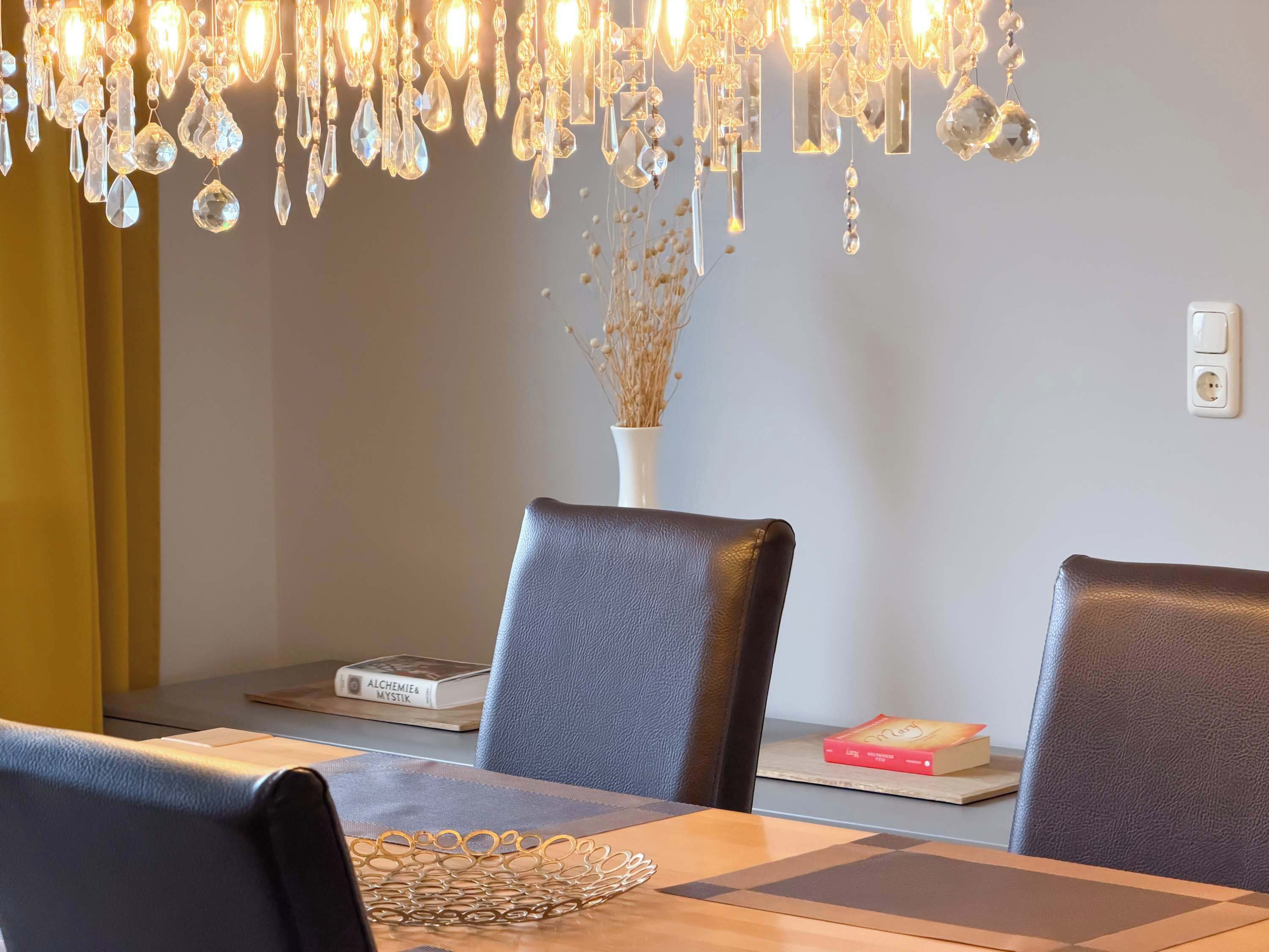 A dining area with a chandelier, a vase with dried plants, and books on the table.