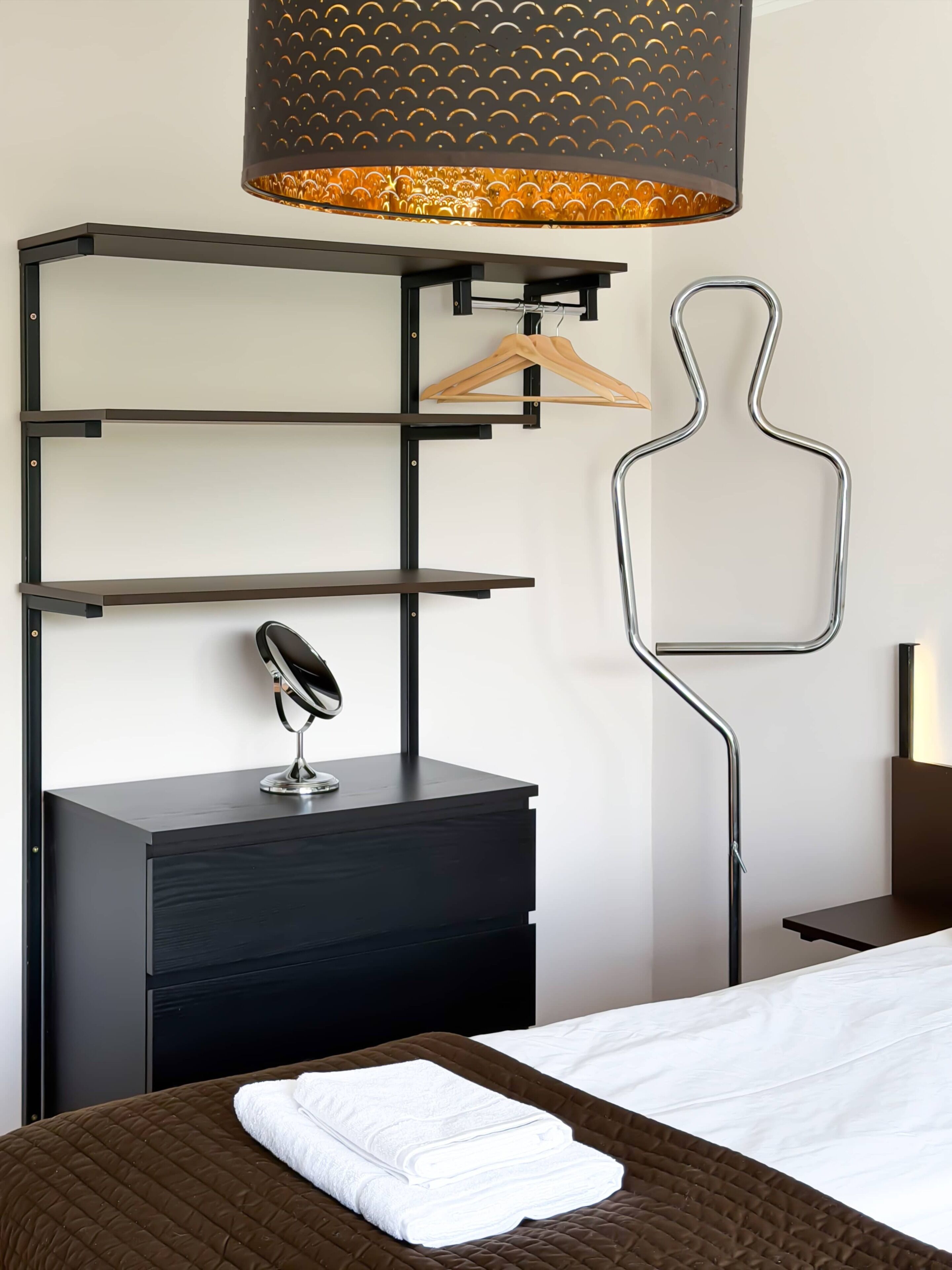 A hotel room with a bed, a bedside table with a lamp, a hanging light fixture, and a wall-mounted shelf with a hanger.
