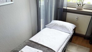 Uma cama de solteiro com manta cinzenta e almofadas brancas, um calendário de parede branco, uma janela com cortinas finas, um radiador e um pequeno vaso de planta no parapeito.