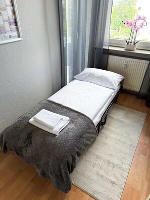 Ein Einzelbett mit grauer Decke und weißen Kissen, ein weißer Wandkalender, ein Fenster mit transparenten Vorhängen, eine Heizung und ein kleiner Topfpflanze auf dem Fensterbrett.