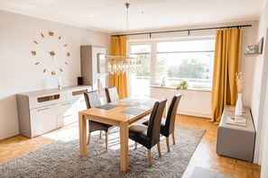 A modern kitchen with a wooden table, chairs, a clock, and a window with curtains.