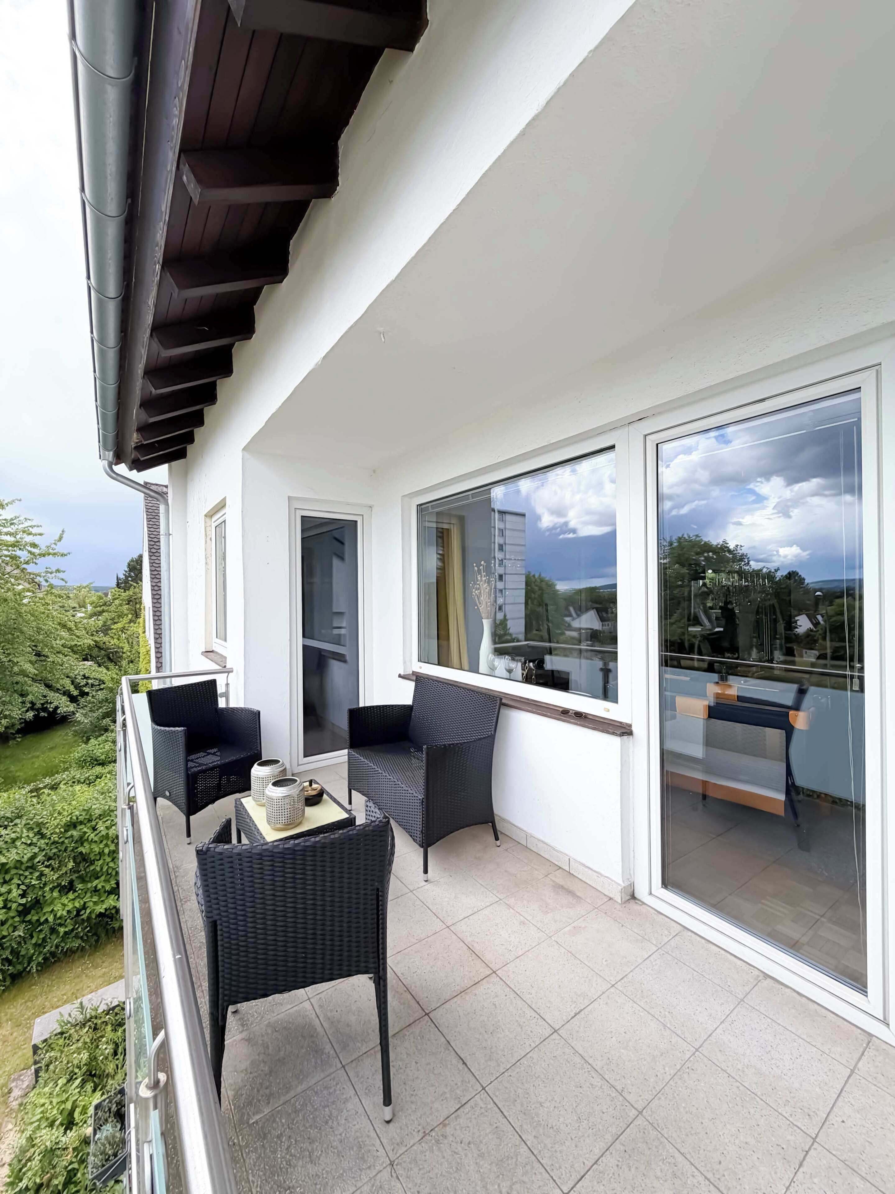 Ein Balkon mit schwarzem Korbmöbelstuhl, einem kleinen Tisch mit einer Vase und Blick auf Grünflächen und Gebäude.