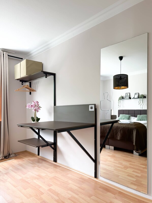 A compact room with a foldable desk, a hanging light fixture, a bed with a headboard, and a small shelf with decorative items.