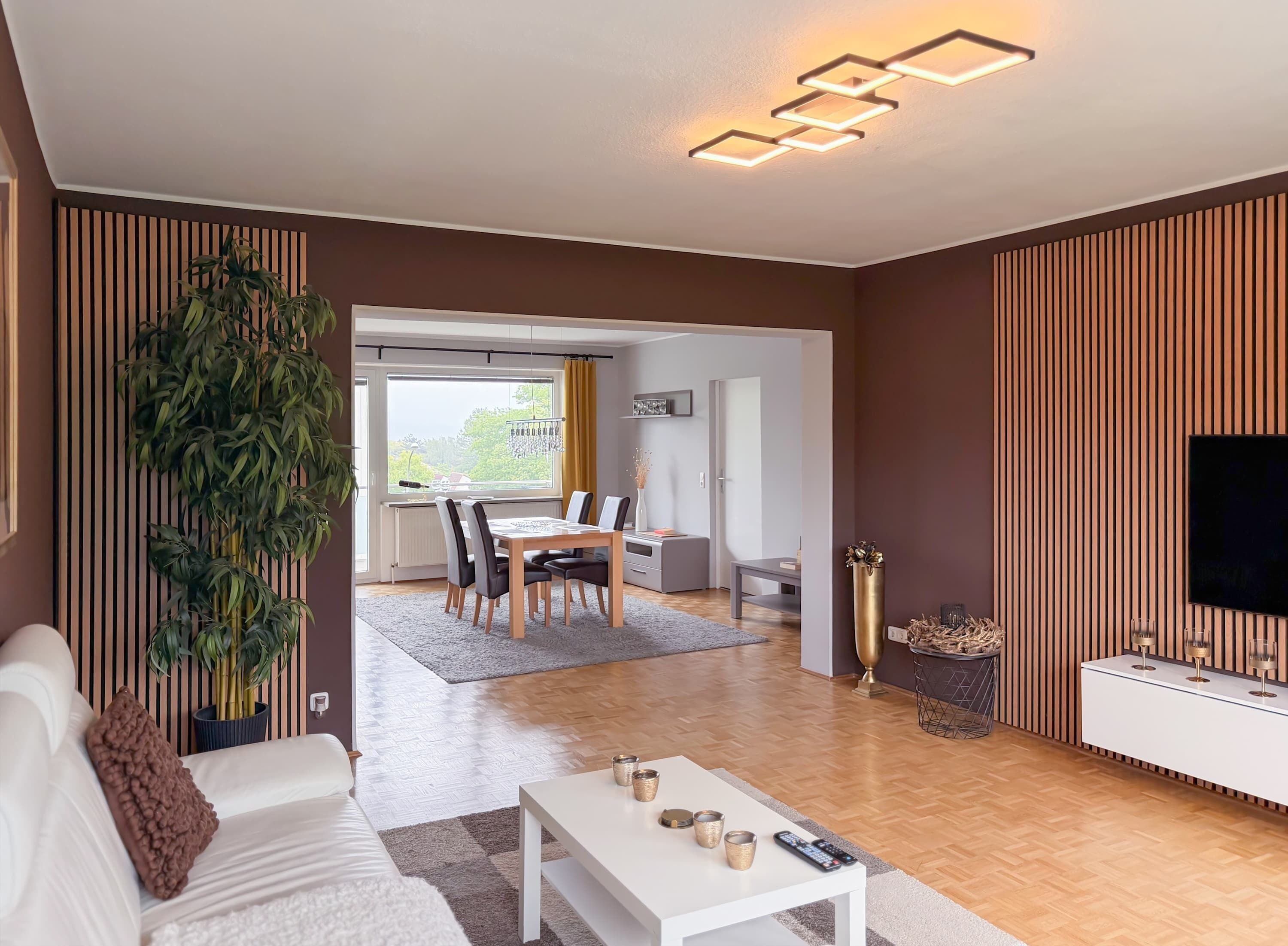 A modern living room with a dining area, a large potted plant, and a flat-screen TV on a wall.