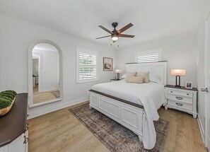 Ein Schlafzimmer mit Bett, Nachttischen, Deckenventilator, Spiegel und Blick in einen anderen Raum.
