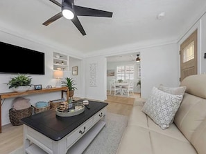 Ein Wohnzimmer mit einer Couch, einem Couchtisch, einem Fernseher, einem Deckenventilator und einem Essbereich im Hintergrund.
