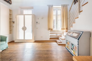 A room with a wooden floor, a green sofa, a white wardrobe, a window with curtains, and a bookshelf with books. - Navigli Garden Terrace - La Vista Apartments (Milano)