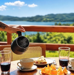 A hand pouring steam from a mug, a table with a plate of food, glasses of red wine, and a scenic view of a lake and mountains.