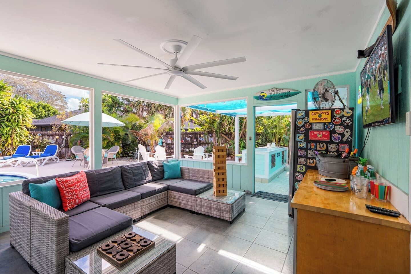 A room with a large sectional sofa, a ceiling fan, a pool area visible through glass doors, and a bar area with a sign reading 'Captain's Saloon'.