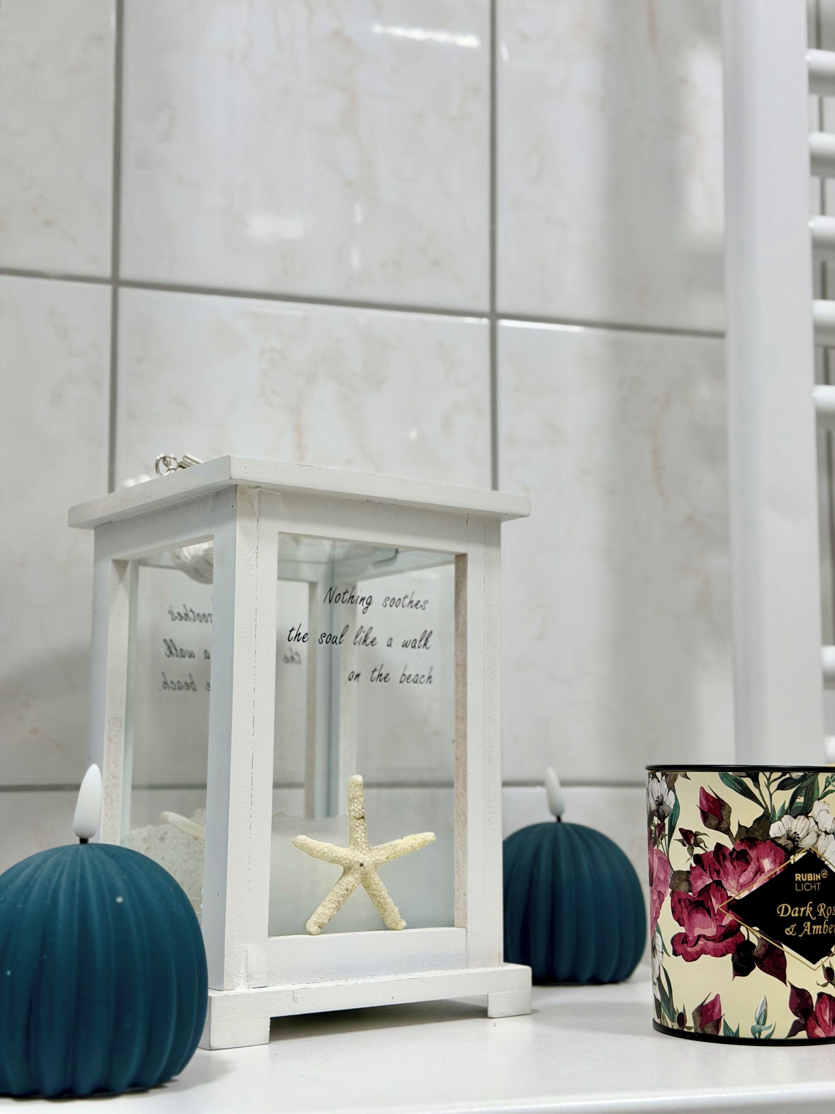 A white lantern with a starfish and a quote, placed on a tiled surface with two blue pumpkins and a floral container.
