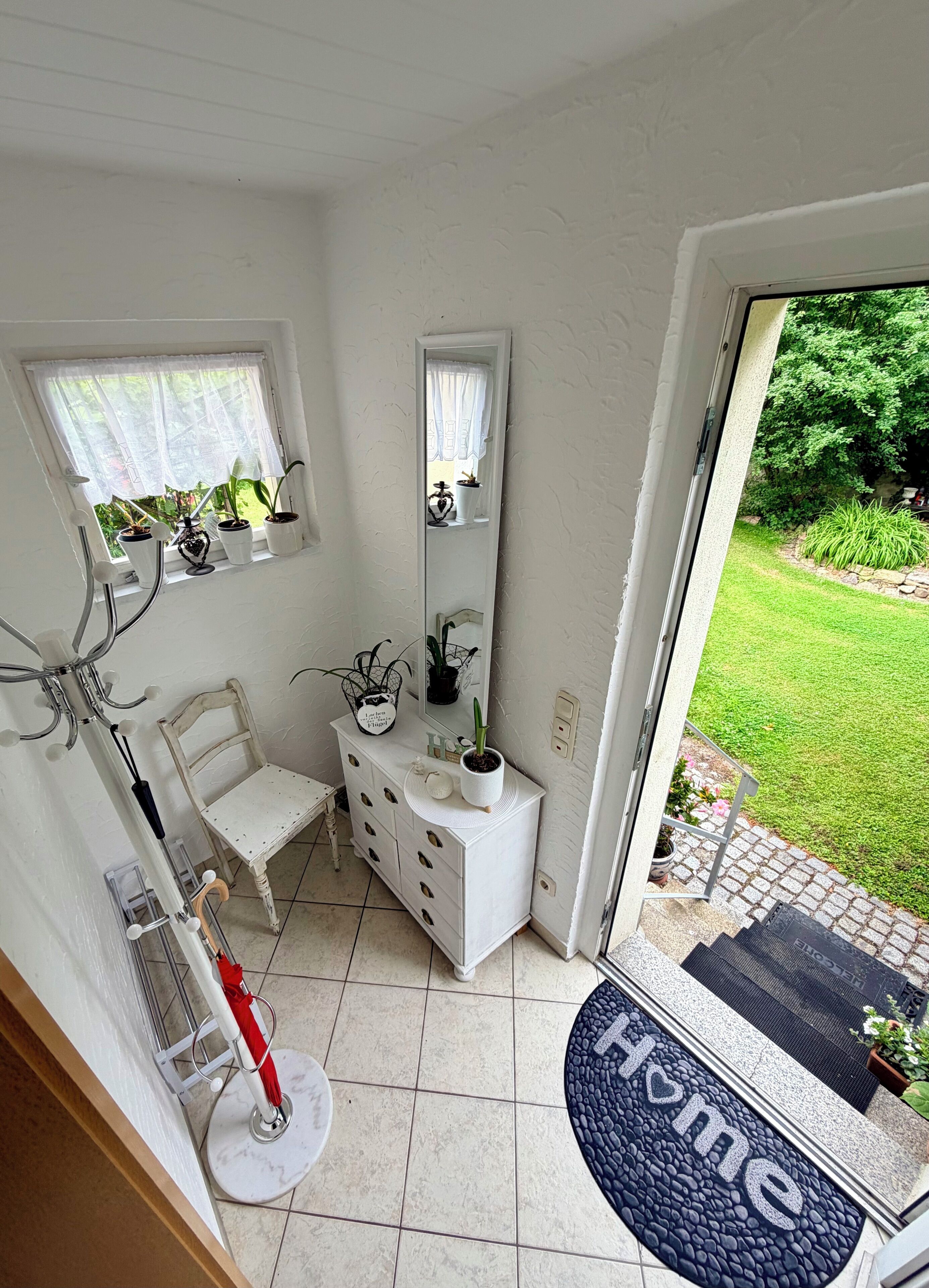 Ein Zimmer mit weißem Schminktisch, einem Sessel, einem Spiegel, einer Topfpflanze und Blick auf einen Garten durch ein Fenster.