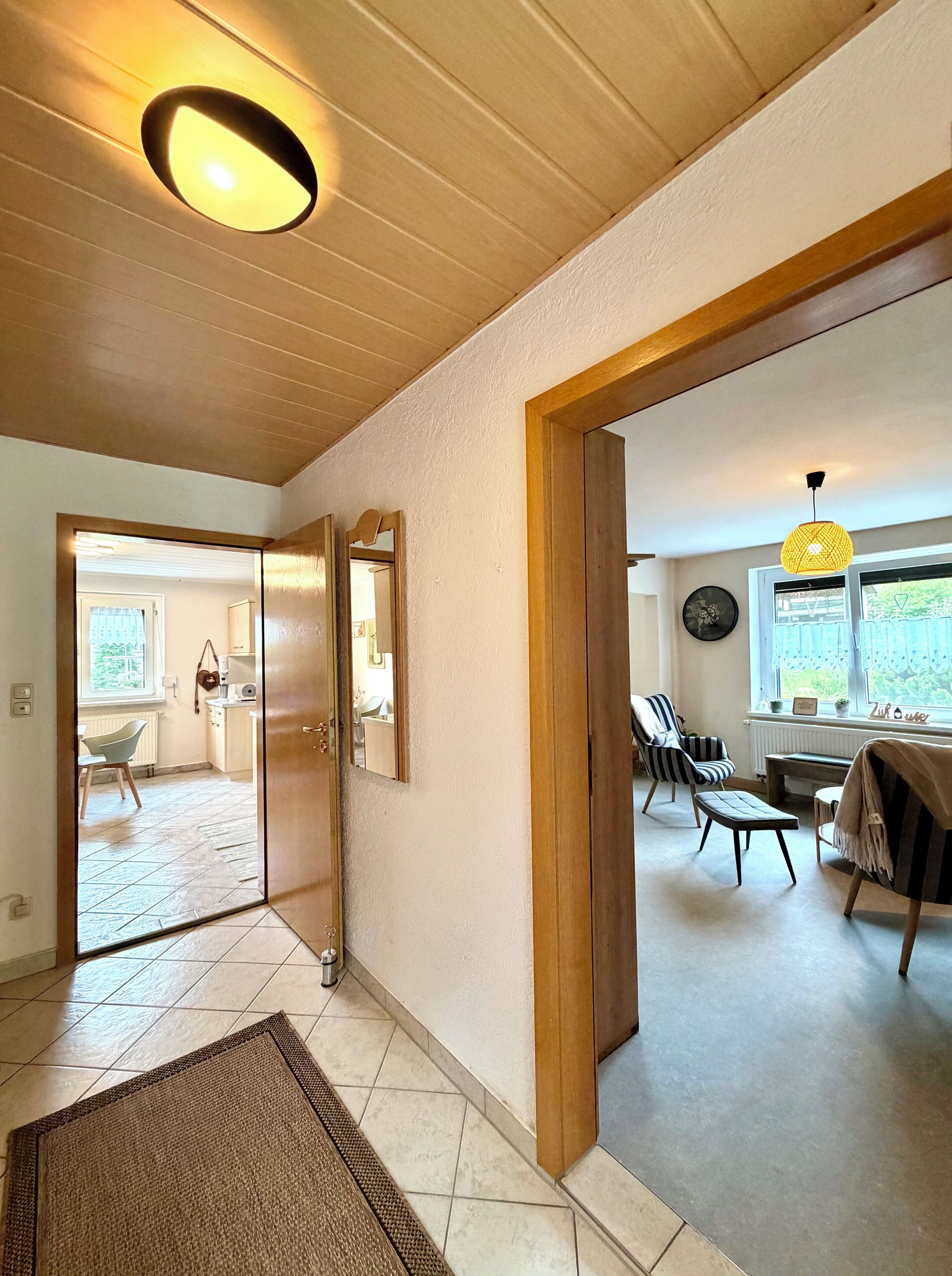 Een ruime woonkamer met houten plafond, grote spiegel, eettafel met stoelen en uitzicht op het landschap door een raam.