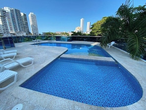 Una piscina en la azotea con agua azul cristalina, rodeada de sillones de descanso y palmeras.