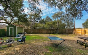 A backyard with a green shed, a trampoline, a fire pit, and a patio table.
