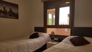 A hotel room with two single beds, a nightstand, a lamp, and a window with a view of buildings. - Apartment Villamartin (Orihuela Costa)