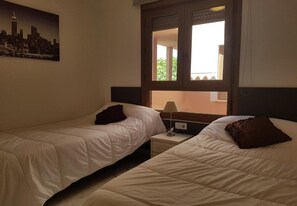 Ein Hotelzimmer mit zwei Einzelbetten, einem Nachttisch, einer Lampe und einem Fenster mit Blick auf Gebäude.