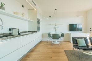 Una cocina moderna con armarios blancos, una zona de comedor con una mesa redonda y sillas, un televisor montado en la pared y suelo de madera.