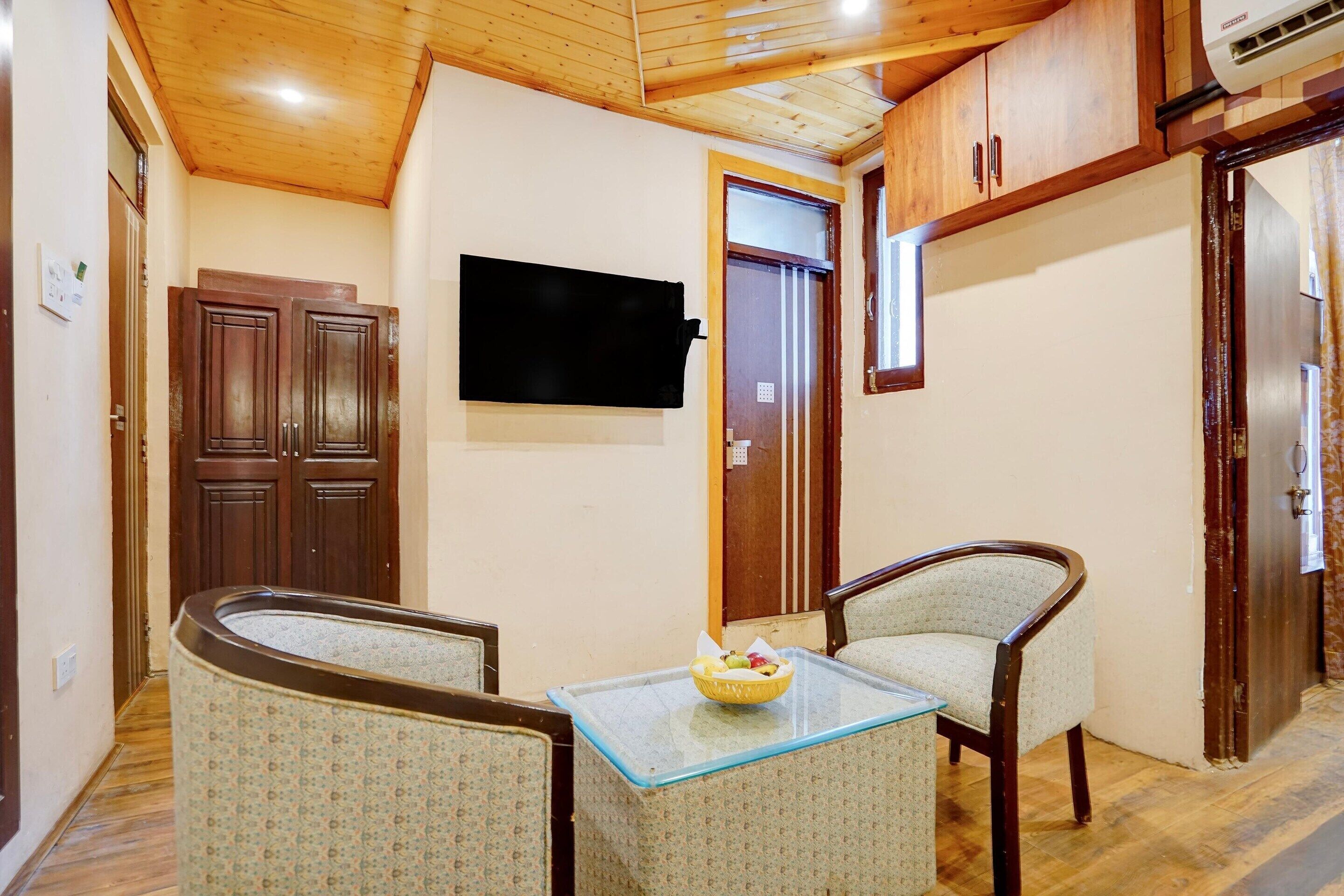 A compact living space with a wooden ceiling, a flat-screen TV mounted on the wall, a small table with a bowl of fruit, and two patterned armchairs.