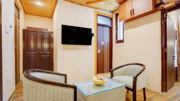 A compact living space with a wooden ceiling, a flat-screen TV mounted on the wall, a small table with a bowl of fruit, and two patterned armchairs.