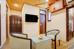 A compact living space with a wooden ceiling, a flat-screen TV mounted on the wall, a small table with a bowl of fruit, and two patterned armchairs. - Saver Double With River View Palette Regency (Anantnag)