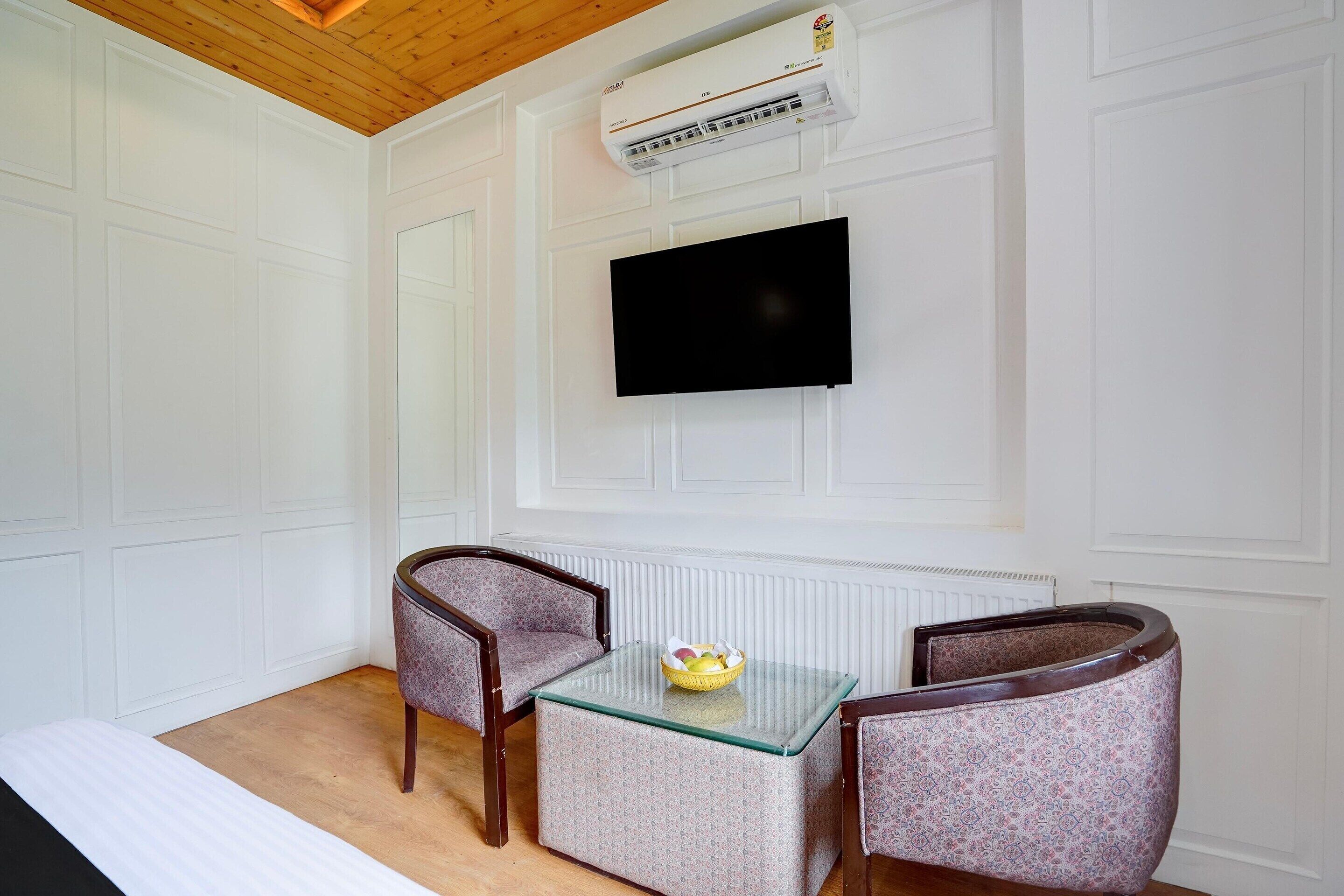 A room with a wooden ceiling, white walls, a mounted flat-screen TV, a glass-top table with chairs, and a radiator.