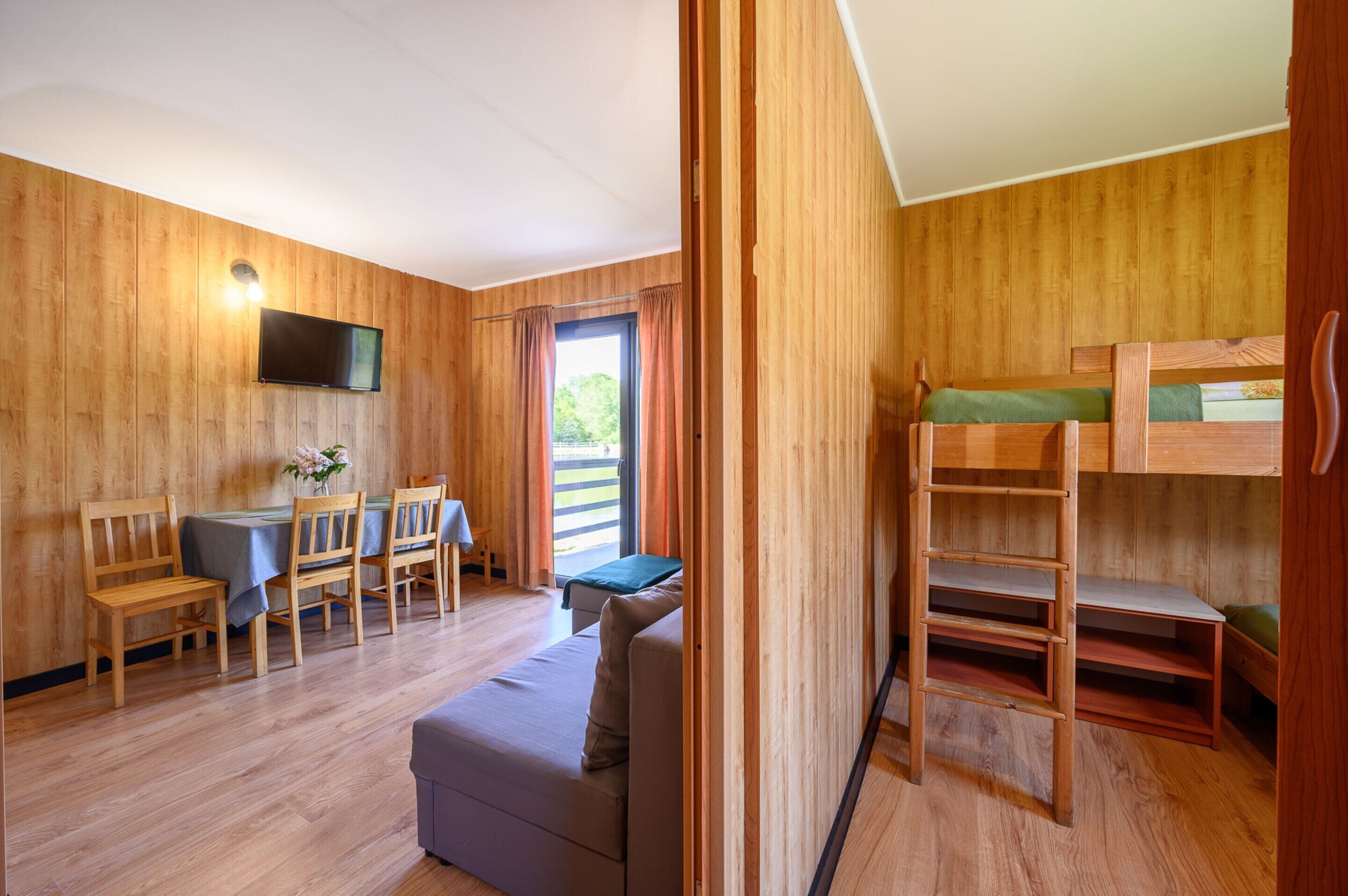 A room with a bunk bed, a sofa, a dining table with chairs, a TV mounted on the wall, and a window with curtains.