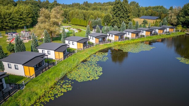 Filas de cabañas modernas junto a un lago tranquilo con lirios acuáticos, rodeadas de exuberante vegetación y un parque infantil.