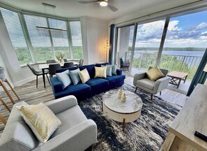 Un salon moderne avec un canapé bleu, des fauteuils gris, une table basse ronde et une vue sur un lac, visible à travers de grandes fenêtres.