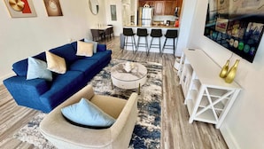 A modern living room with a blue sofa, beige armchair, and a coffee table. There is a kitchen area with bar stools and a TV mounted on the wall. - Balcony Lake Views |  Pool, Gym, WiFi & Parking (Orlando)