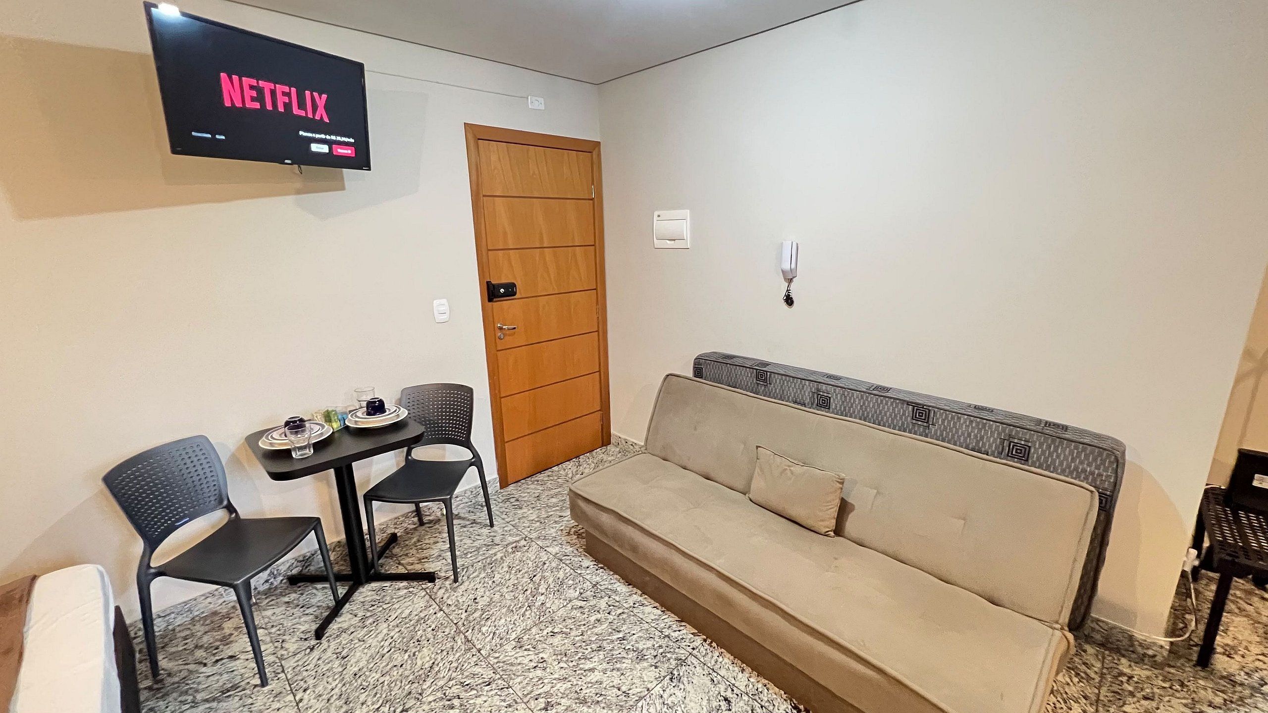 Una stanza piccola con un divano, due poltrone, un tavolo, una televisione che mostra 'NETFLIX' e una porta.