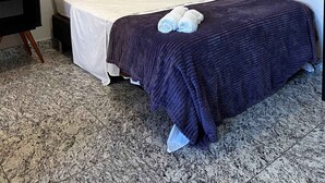 En säng med en mörkblå filt, en vit kudde och två hoprullade handdukar vid fotändan. På väggen ovanför sängen hänger en inramad tavla.