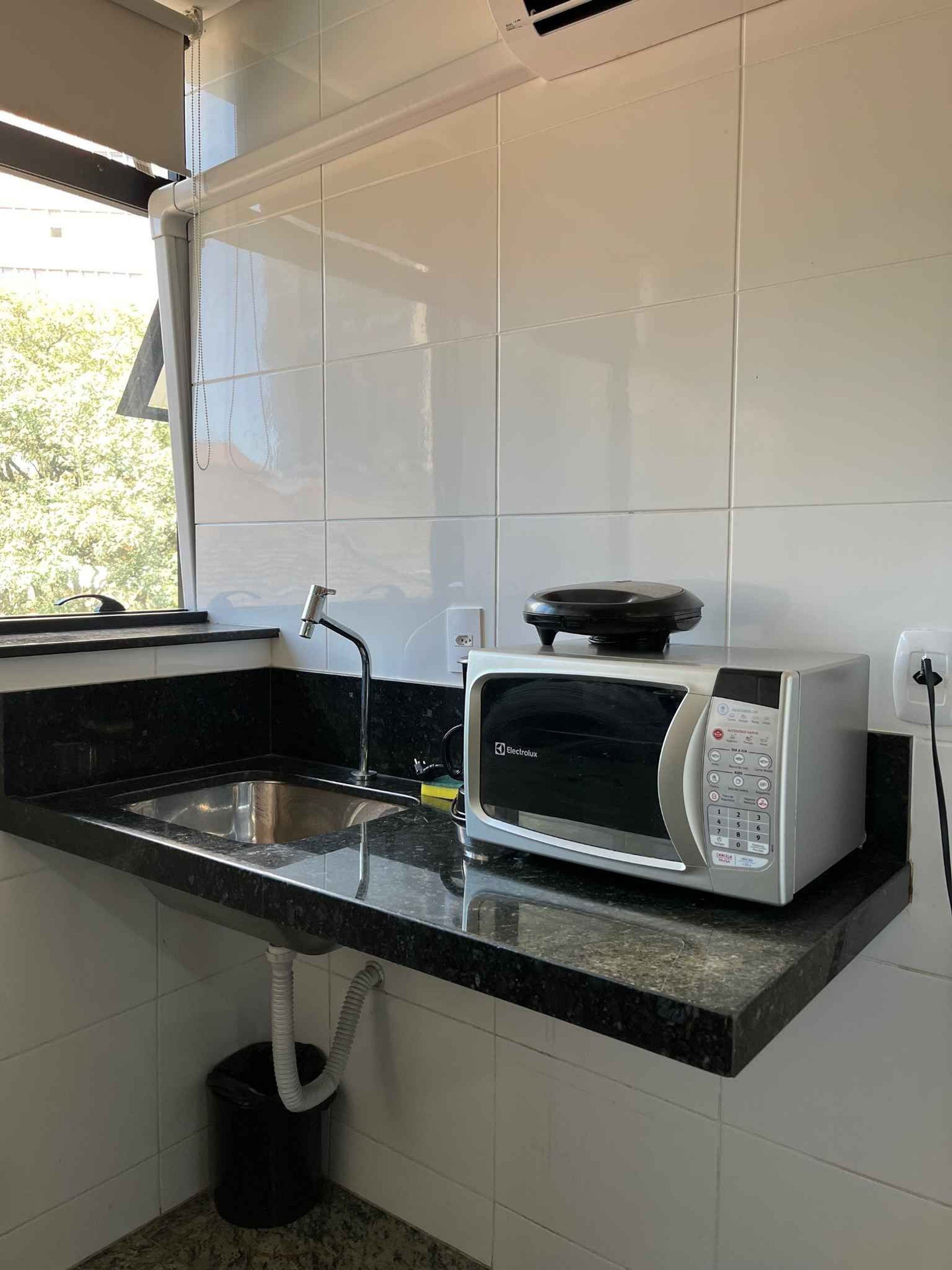Una cocina con microondas integrado, un lavamanos de acero inoxidable y una ventana con persianas.