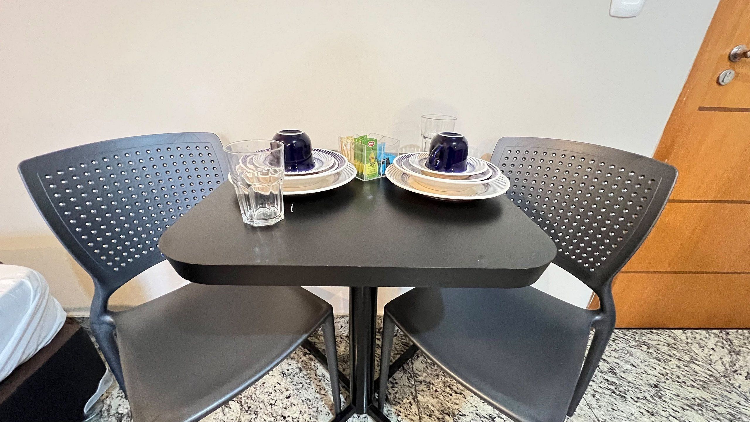 A small table with two chairs, a glass, a blue cup, and a plate with a blue cup on it.