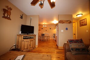 Ein Wohnzimmer mit Fernseher auf einem Holzschrank, eine Essecke mit Tisch und Stühlen sowie eine Küche mit Kühlschrank und Herd.