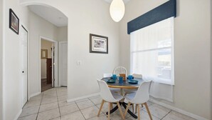 Un coin repas avec une table dressée pour quatre personnes, des chaises blanches, un luminaire suspendu et une fenêtre avec des stores.