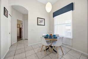 A dining area with a table set for four, white chairs, a hanging light fixture, and a window with blinds. - Dreamers Retreat! Spacious Disney-Area Pool Home for Families & Adult Getaways (Kissimmee)