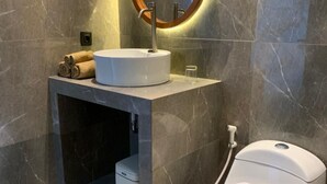 A modern bathroom with a round mirror, a white sink, a toilet, and a shower. - Cozzy Sagas  House Tabanan (Marga)