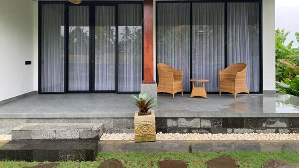 A covered outdoor seating area with two wicker chairs and a small table, a potted plant, and a stone pathway leading to a building with large sliding doors. - Cozzy Sagas  House Tabanan (Marga)